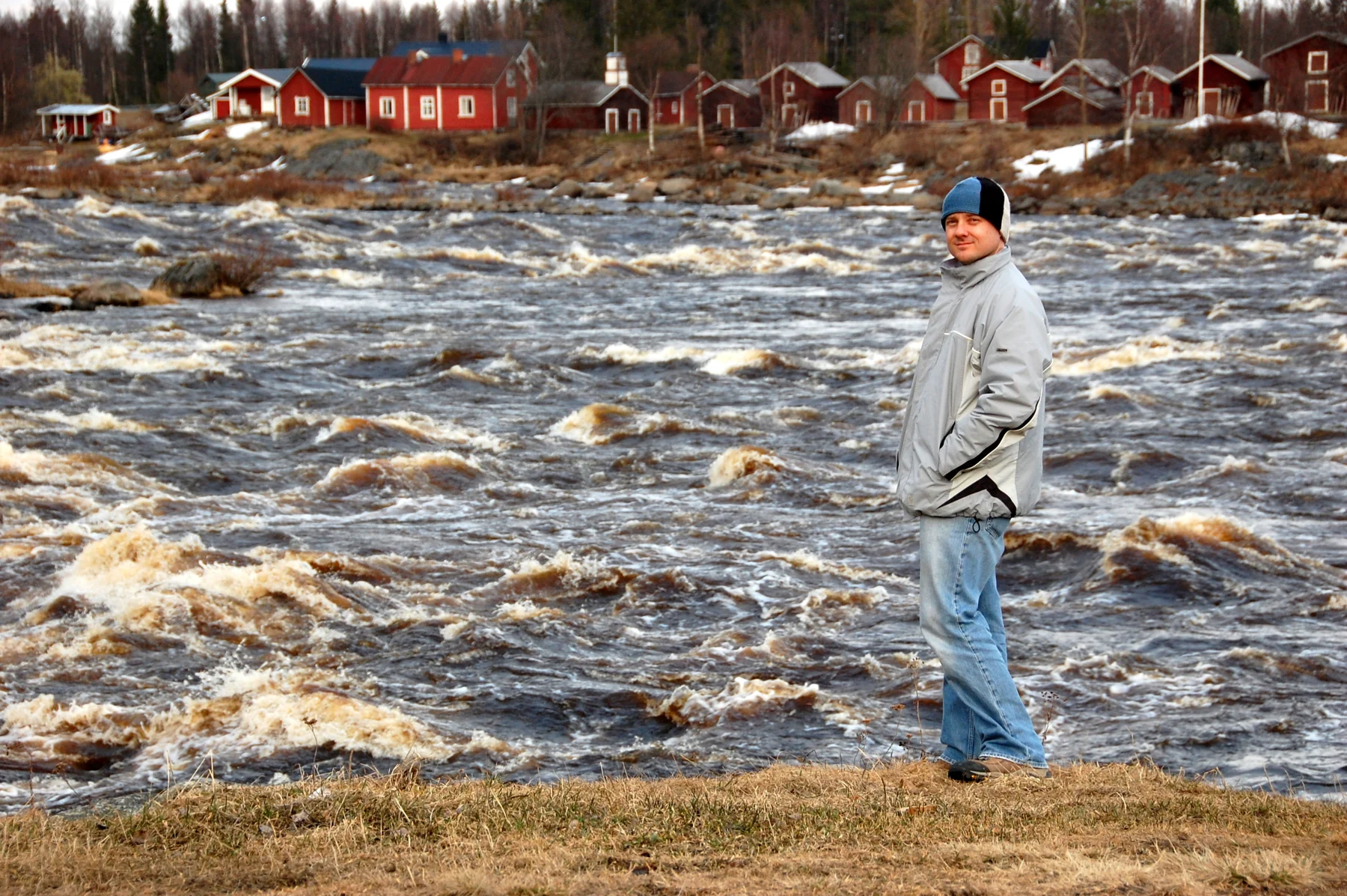 A family member beside the rushing Torne River, with red wooden buildings across the water